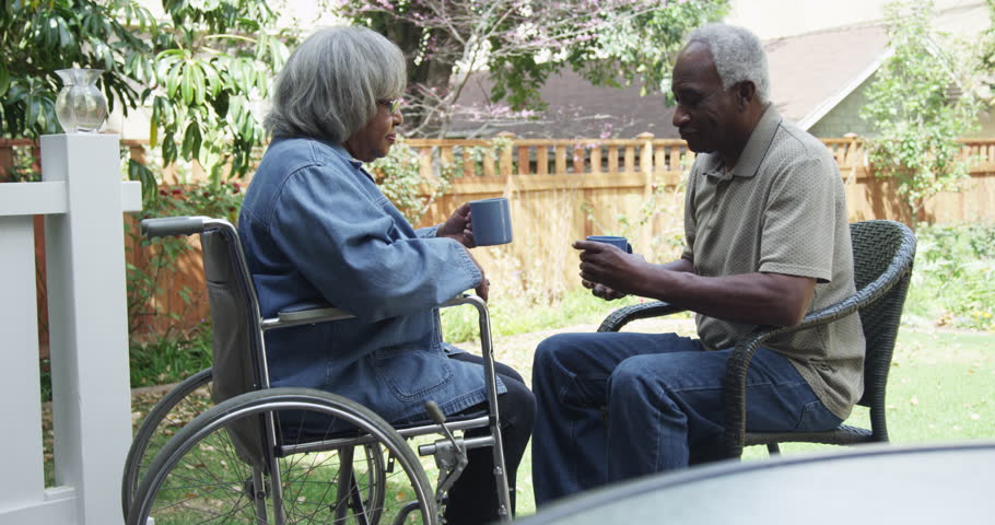 An older man seated in a chair speaks with an older woman in a wheelchair.