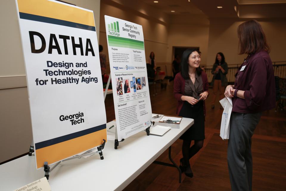 Two people talk next to a table with signage about design and technology for healthy aging.
